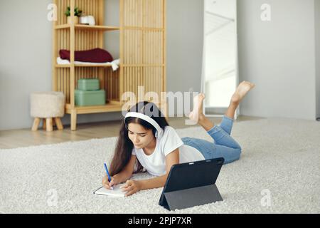 Jolie pré-adolescent 12s fille porter des écouteurs sans fil, couché sur le sol écouter cours audio étudier, à la maison école utiliser la tablette numérique. Utilisation des technologies modernes f Banque D'Images