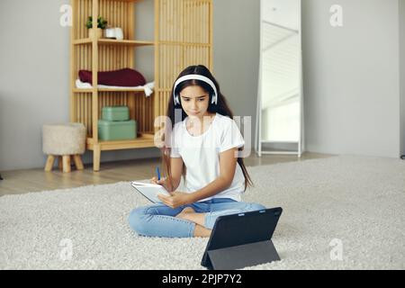 Jolie pré-adolescent 12s fille portent des écouteurs sans fil, assis sur le sol écouter cours audio étudiant, école à domicile à l'aide de la tablette numérique. Utilisation des technologies modernes f Banque D'Images