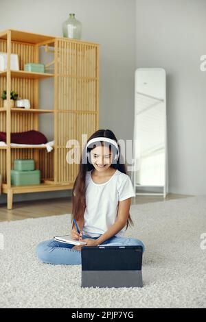 Jolie pré-adolescent 12s fille portent des écouteurs sans fil, assis sur le sol écouter cours audio étudiant, école à domicile à l'aide de la tablette numérique. Utilisation des technologies modernes f Banque D'Images