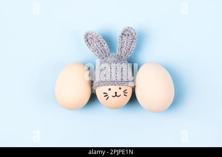 Œufs de Pâques aux visages amusants et chapeaux crochetés gris avec oreilles de lapin sur fond bleu. Concept joyeuses Pâques. Carte de vœux. Banque D'Images