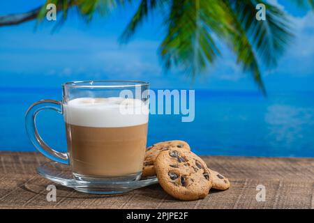 Tasse en verre de cappuccino chaud avec biscuits sur une table en bois avec eau de mer, cococotier et fond bleu ciel le jour ensoleillé d'été en métro Banque D'Images