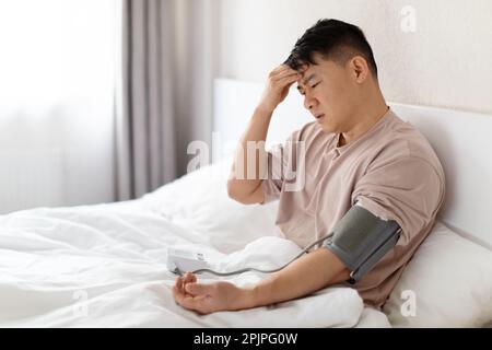 Homme asiatique portant un pyjama assis dans le lit, vérifier la tension artérielle Banque D'Images