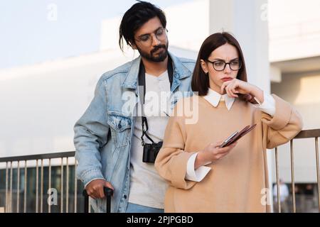 Malheureux pensif jeune indien et femme européenne dans une ambiance décontractée et des lunettes avec téléphone, passeport et billets Banque D'Images