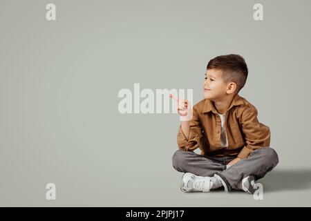 Souriant caucasien 6 ans petit garçon dans une ambiance décontractée s'asseoir sur le sol, en pointant le doigt sur l'espace de copie Banque D'Images