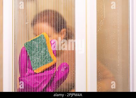 La main d'une femme dans des gants en latex tient une éponge et lave les parois en verre de la cabine de douche dans la salle de bains. Mise au point sélective, espace de copie. Banque D'Images