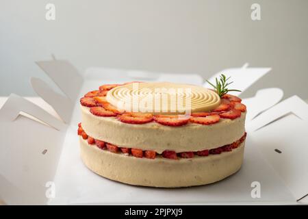 Gâteau d'anniversaire végétalien à la vanille et à la fraise recouvert de chocolat blanc dans une boîte ouverte en carton sur une table et un fond blanc Banque D'Images