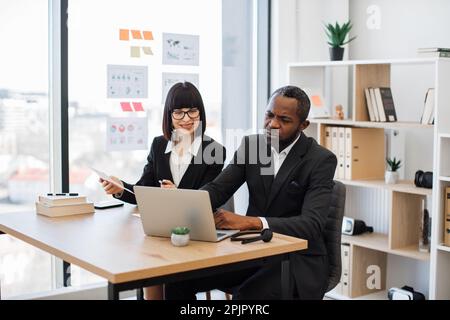 Un homme d'affaires africain concentré et son agréable assistant caucasien analysant des données sur un ordinateur portable moderne tout en étant assis dans un lieu de travail créatif. Collègues d'affaires discutant du nouveau projet financier au bureau. Banque D'Images