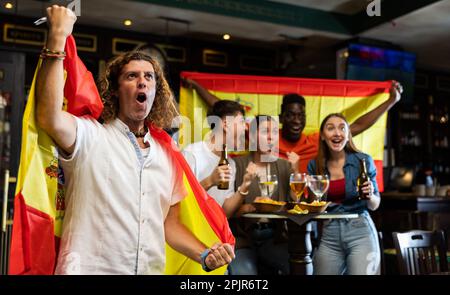 Des fans de sport très divers avec drapeau espagnol se réjouissent du jeu gagnant avec des verres de bière et de frites au pub Banque D'Images