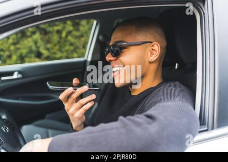 Jeune adulte habillé à la mode Noir homme dans des lunettes de soleil assis derrière une roue et parlant par haut-parleur sur son téléphone. Portrait pris par la vitre conducteur. Photo de haute qualité Banque D'Images