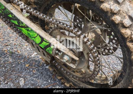 Chaîne et pneu arrière noir à boutons en caoutchouc recouvert de boue séchée sur un vélo de terre stationné sur une surface de gravier. Banque D'Images