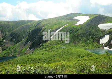 Un petit lac sur une colline escarpée et de hautes roches avec des résidus de neige sur des pentes herbeuses sous un ciel nuageux d'été. Lacs Ivnovskie, Khakassia, Sibérie, Rus Banque D'Images