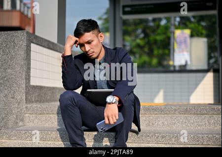 Un homme d'affaires asiatique millénaire fatigué et stressé est assis sur un escalier devant la station de métro dans une ville par beau temps. échec du travail, obtention Banque D'Images