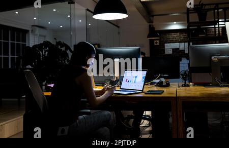 Femme d'affaires asiatique assise au bureau, utilisant un ordinateur portable et un smartphone, travaillant tard au bureau Banque D'Images