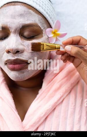 Femme afro-américaine décontractée de grande taille ayant un masque de beauté appliqué au visage au spa Banque D'Images