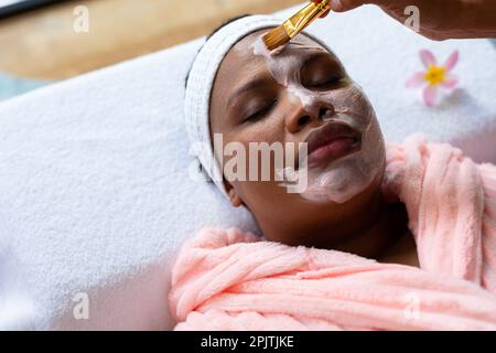 Femme afro-américaine décontractée de grande taille ayant un masque de beauté appliqué au visage au spa Banque D'Images