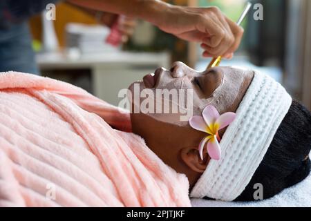 Femme afro-américaine décontractée de grande taille ayant un masque de beauté appliqué au visage au spa Banque D'Images