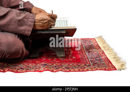 Homme musulman assis et lisant le Coran sur le tapis de prière isolé sur fond blanc Banque D'Images