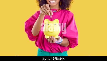 Un enfant afro-américain heureux qui économise de l'argent met de la pièce dans une petite banque de pigey Banque D'Images