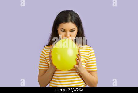 Une femme gaie avec une drôle d'expression faciale gonfle ballon sur fond violet pastel. Banque D'Images