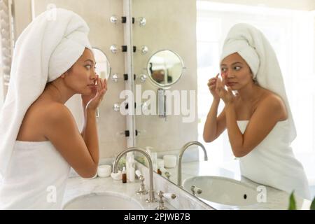 Bonne femme asiatique portant une serviette sur la tête regardant dans le miroir de salle de bains appliquant sous les taches de beauté des yeux Banque D'Images