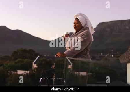 Femme afro-américaine de bonne taille, portant un peignoir et une serviette sur la tête, buvant du thé sur le balcon Banque D'Images