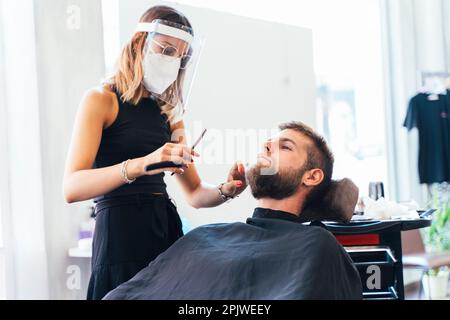 Coiffeur avec masque facial de protection se raser un homme dans un salon Banque D'Images