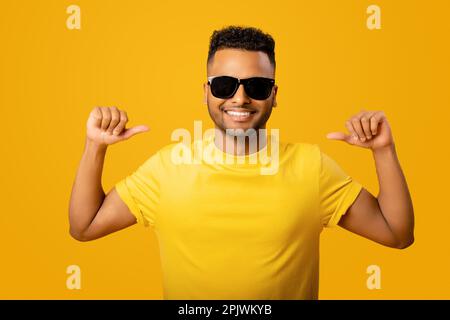 Souriant gai jeune indien dans des lunettes de soleil et t-shirt isolé sur fond jaune pointant vers lui-même, individu-confiant ethnique de choix, la publicité Banque D'Images