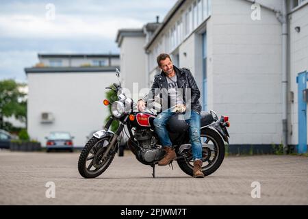 Portrait d'un homme adulte souriant vêtu d'une veste en cuir et d'un Jean bleu sur une moto. Un homme pose sur une moto rétro garée. Banque D'Images