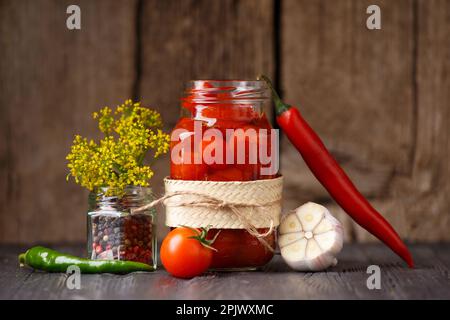 Tomates cerises marinées dans un pot en verre ouvert, piments, ail, assaisonnements, épices et herbes pour réaliser un gros plan de la marinade sur fond de bois sombre. Banque D'Images