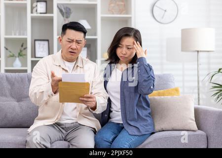 Contrarié famille asiatique, homme et femme assis ensemble à la maison dans le salon sur le canapé, couple a reçu lettre notification enveloppe avec de mauvaises nouvelles, malheureux avec le message reçu. Banque D'Images