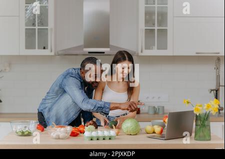 Un couple marié cuisiniers ensemble à l'aide d'un ordinateur portable, à la recherche de recettes sur l'Internet. Un homme afro-américain et sa femme préparent une salade dans le kit Banque D'Images