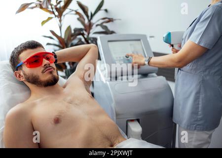 Jeune homme recevant une épilation photo lors d'une visite du centre de beauté dans un salon, esthéticienne professionnelle effectuant une procédure d'épilation laser sur le client Banque D'Images