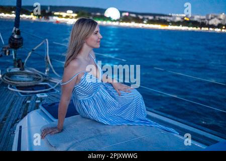 La jeune femme en robe bleue et blanche est assise à la poupe du bateau. Blonde fille regarde les lumières de la ville de nuit. Promenade en soirée sur le bateau le long de l' Banque D'Images