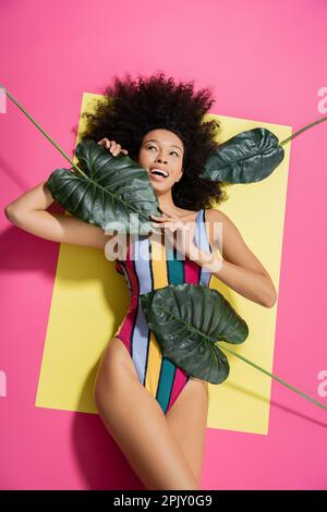 vue de dessus de la femme afro-américaine excitée dans un maillot de bain coloré faisant bronzer près des feuilles de palmier vertes sur le rose Banque D'Images