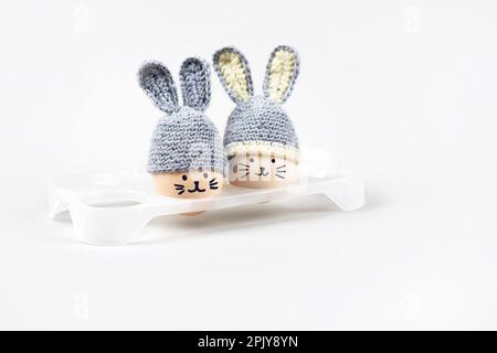 Deux œufs de Pâques aux visages amusants et chapeaux crochettés gris aux oreilles de lapin dans un support d'œuf blanc sur fond gris. Concept joyeuses Pâques. Accueil voiture Banque D'Images