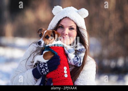 Jeune femme en veste d'hiver tenant son terrier Jack Russell sur les mains - arrière-plan d'arbres flous Banque D'Images