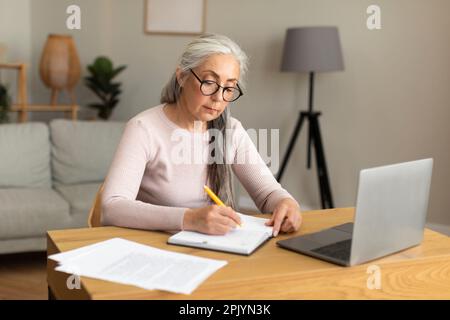 Concentré sérieuse occupé jolie vieille dame caucasienne dans les verres fait des notes à la table avec l'ordinateur Banque D'Images