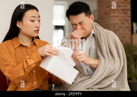 Homme asiatique d'âge moyen soufflant le nez dans les serviettes, épouse affectueuse traitant son mari malade, prenant soin Banque D'Images