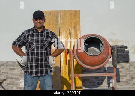 Image d'un ouvrier de construction souriant avec un mélangeur de béton et des outils de construction en arrière-plan sur un chantier de construction. Travail manuel et profession Banque D'Images