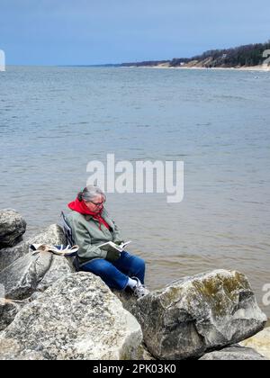 Femme portant un manteau et lisant un livre sur des rochers de plage Banque D'Images