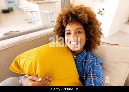 Portrait de jeune femme biracial souriante avec cheveux afro relaxant avec coussin jaune sur le canapé Banque D'Images