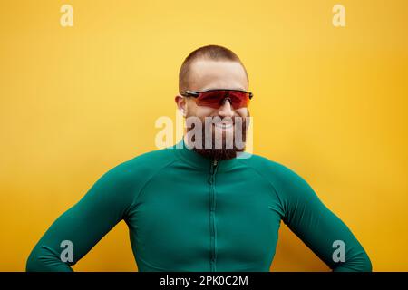 Gros plan d'un jeune homme européen heureux et séduisant avec une barbe portant des lunettes et des vêtements de sport verts regardant l'appareil photo sur un mur jaune. Banque D'Images