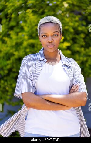 Jeune femme afro-américaine aux cheveux courts posant avec les bras croisés contre les plantes dans la cour Banque D'Images