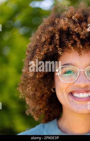 Petit visage de jeune femme biracial avec des cheveux afro souriant et regardant l'appareil photo dans la cour Banque D'Images