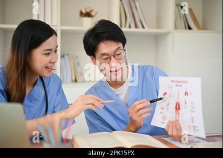 Un jeune étudiant en médecine asiatique intelligent explique et donne des cours de tutorat sur le système du corps humain à son ami dans une bibliothèque. Banque D'Images