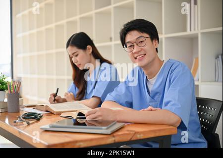 Jeune homme asiatique beau et intelligent étudiant en médecine dans un uniforme étudiant avec son ami dans un espace de travail commun de bibliothèque. Banque D'Images