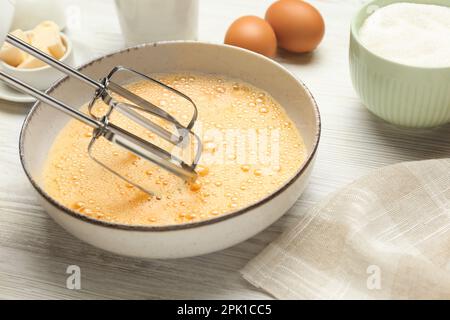 Battre les œufs dans un bol avec un batteur sur une table en bois blanc, gros plan Banque D'Images