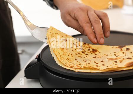 Homme cuisant une délicieuse crêpe sur une machine à crêpes électrique à table, en gros plan Banque D'Images