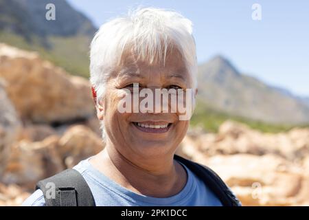 Portrait d'une femme biraciale âgée heureuse portant un sac à dos, regardant l'appareil photo et souriant dans les montagnes Banque D'Images