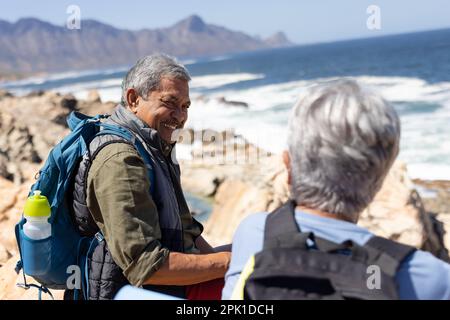 Heureux couple biracial senior assis sur des rochers au bord de la mer, souriant Banque D'Images